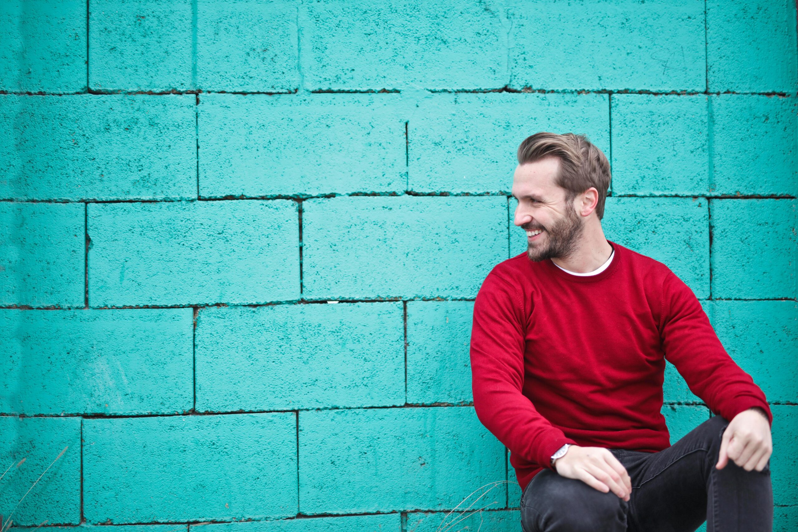 A cheerful man in a red sweater sits against a turquoise wall, enjoying a moment of happiness and laughter.
