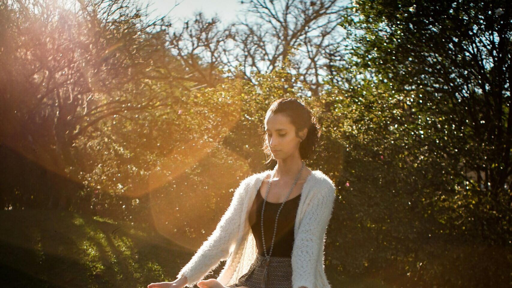 Woman in tranquil meditation pose outdoors, bathed in sunlight, surrounded by lush greenery.