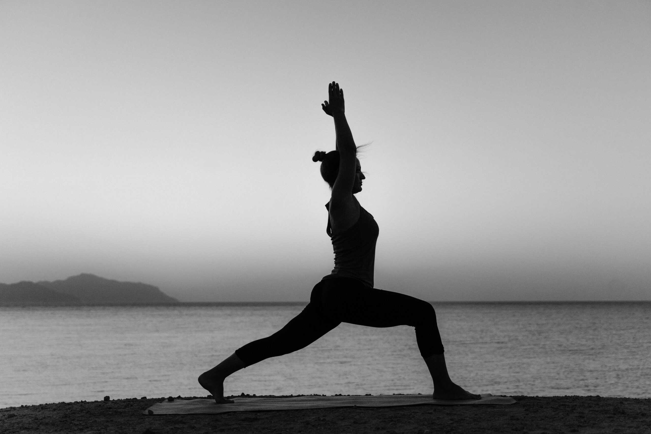 Grayscale silhouette of a woman practicing yoga on a beach at sunset.